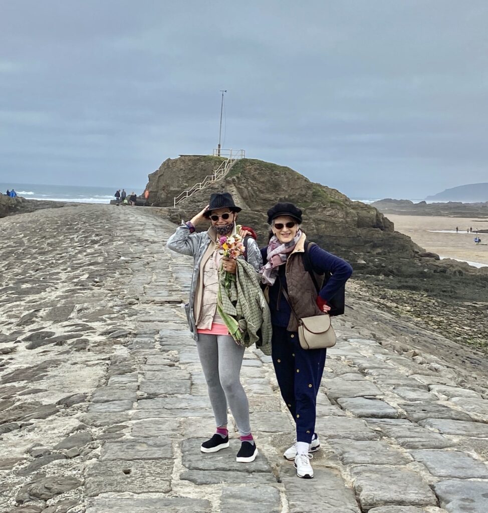 Two white women posing together on a cobbled pathway in front of the coast.